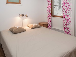 Ein Schlafzimmer mit einem Bett, Kissen, einem Nachttisch, einer Lampe und einem Fenster mit Vorhängen.