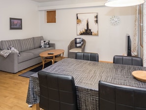 Ein möbliertes Wohnzimmer mit einer Couch, einem Esstisch mit Stühlen, einem kleinen runden Tisch, einem Fernseher und einem Bild an der Wand.