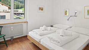 Ein Hotelzimmer mit zwei Betten, Blick auf ein Gebäude und einen Wald durch ein Fenster, einem Heizkörper und einem grünen Stuhl.