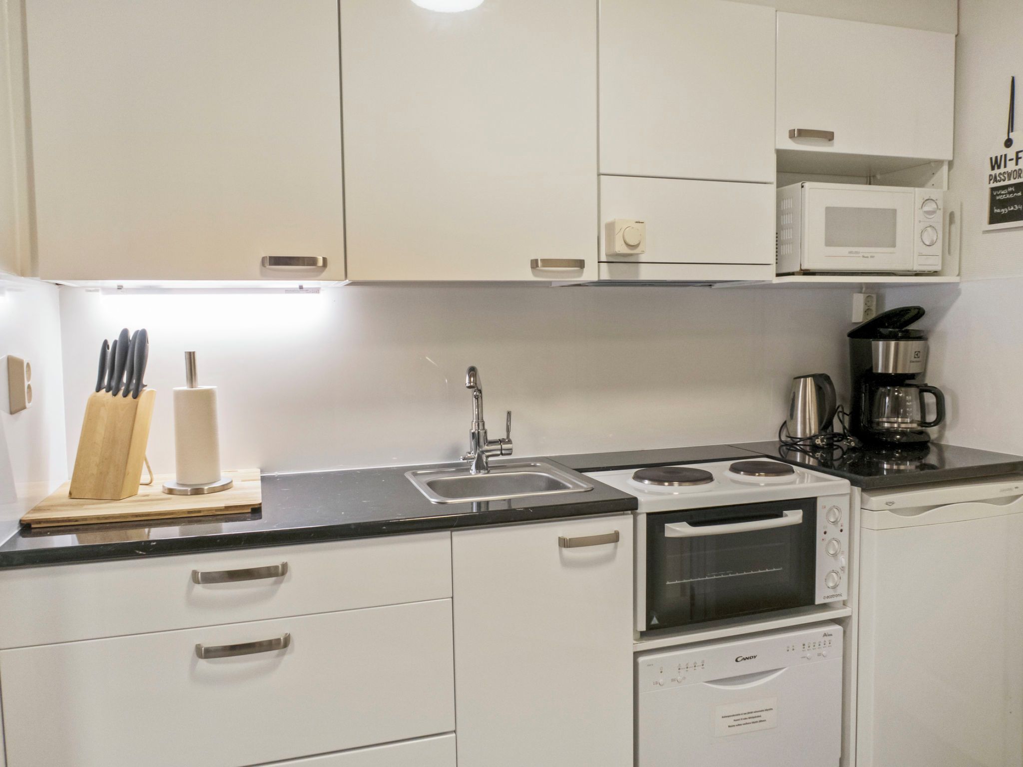 Une cuisine de taille réduite, avec des meubles blancs, un évier, une cuisinière, un four à micro-ondes et une machine à café.