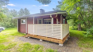 En hyggelig hytte med træterrasse, omgivet af træer og en grussti.