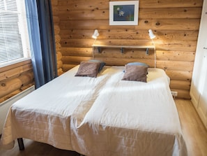 Cama doble con dos almohadas, cabecera de madera, un cuadro enmarcado sobre la cama y una mesita de noche con una lámpara.