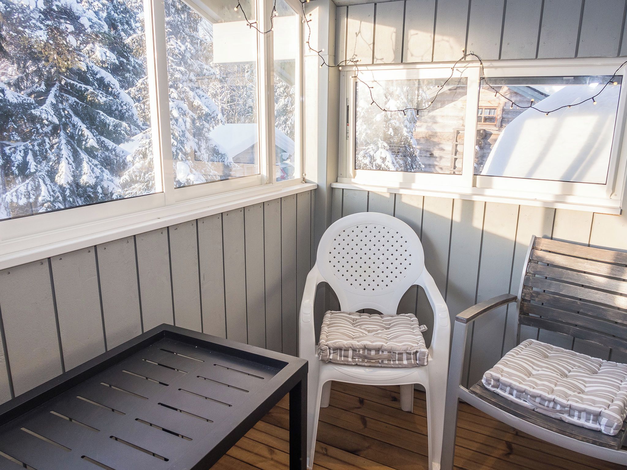 A corner room with a window overlooking a snowy landscape, a white plastic chair with a cushion, and a wooden bench.