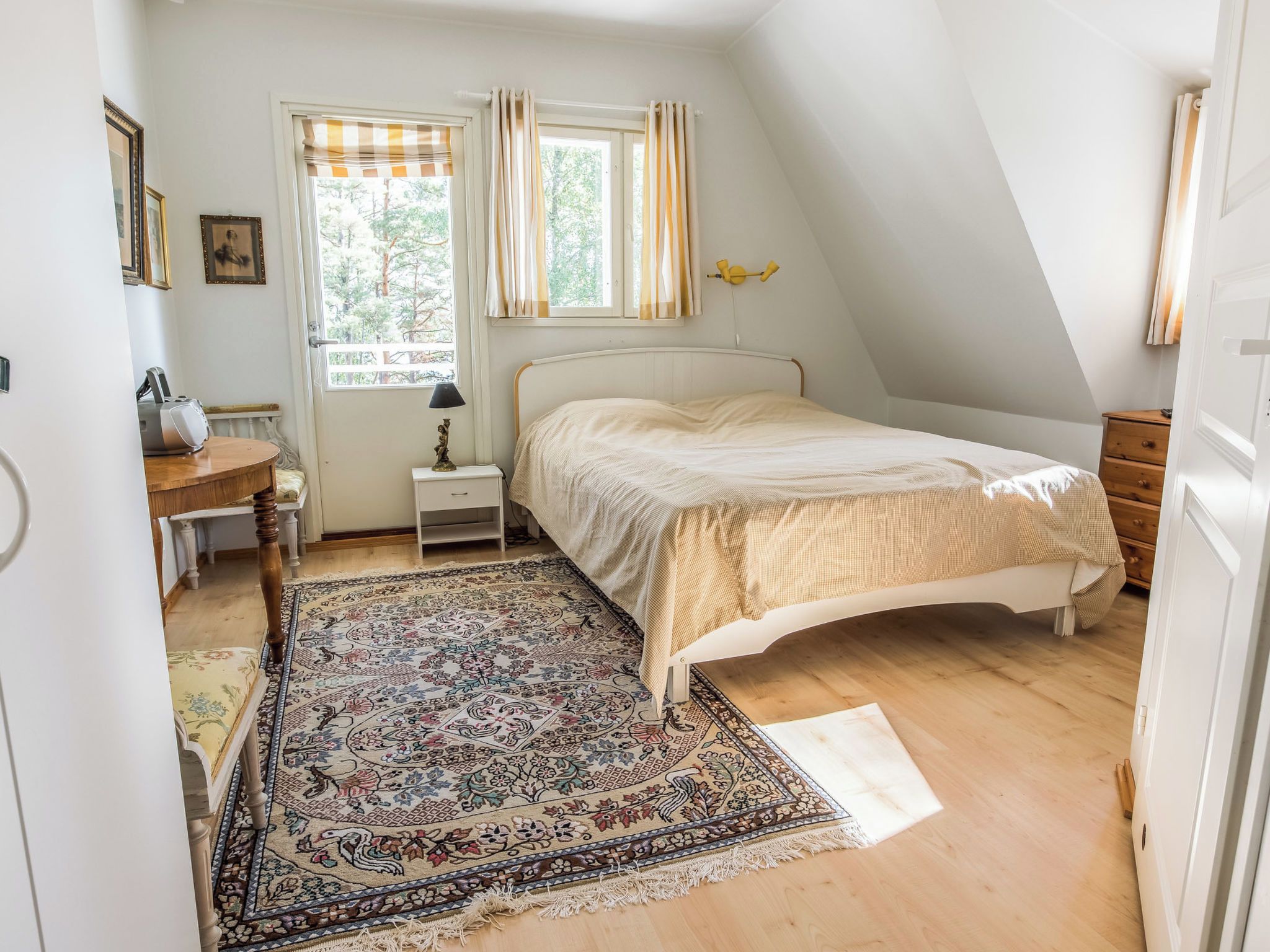 Ein Schlafzimmer mit einem Bett, einem Nachttisch, einer Lampe, einem Fenster mit Vorhängen, einer Tür und einem gemusterten Teppich.
