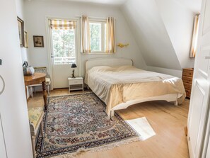 Een slaapkamer met een bed, nachtkastje, lamp, raam met gordijnen, een deur en een kamerbreed tapijt.