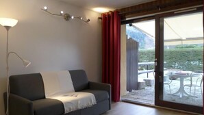Un salon moderne avec un canapé gris, une lampe sur pied et une porte-fenêtre coulissante donnant sur une terrasse extérieure équipée d’une table et de chaises.