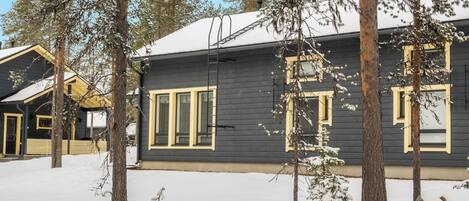 A cabin with a dark exterior and yellow trim, surrounded by snow-covered ground and trees.