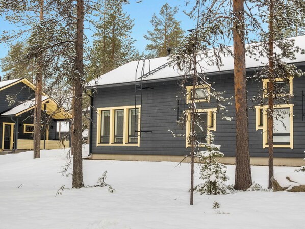A cabin with a dark exterior and yellow trim, surrounded by snow-covered ground and trees.