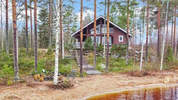 Een chalet met een houten terras en een trap naar een tweede verdieping, omringd door bos en een waterpartij.