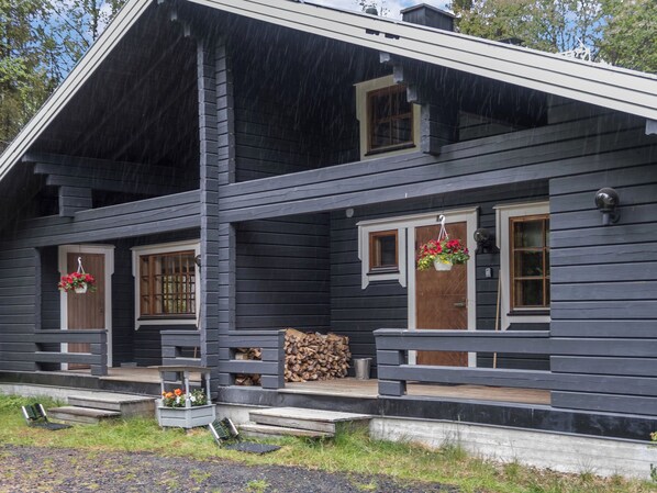 A two-story log cabin with a porch, a firewood stack, and flower baskets.