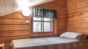 A small, cozy cabin room with a single bed, a window with a curtain, and a wooden ceiling.
