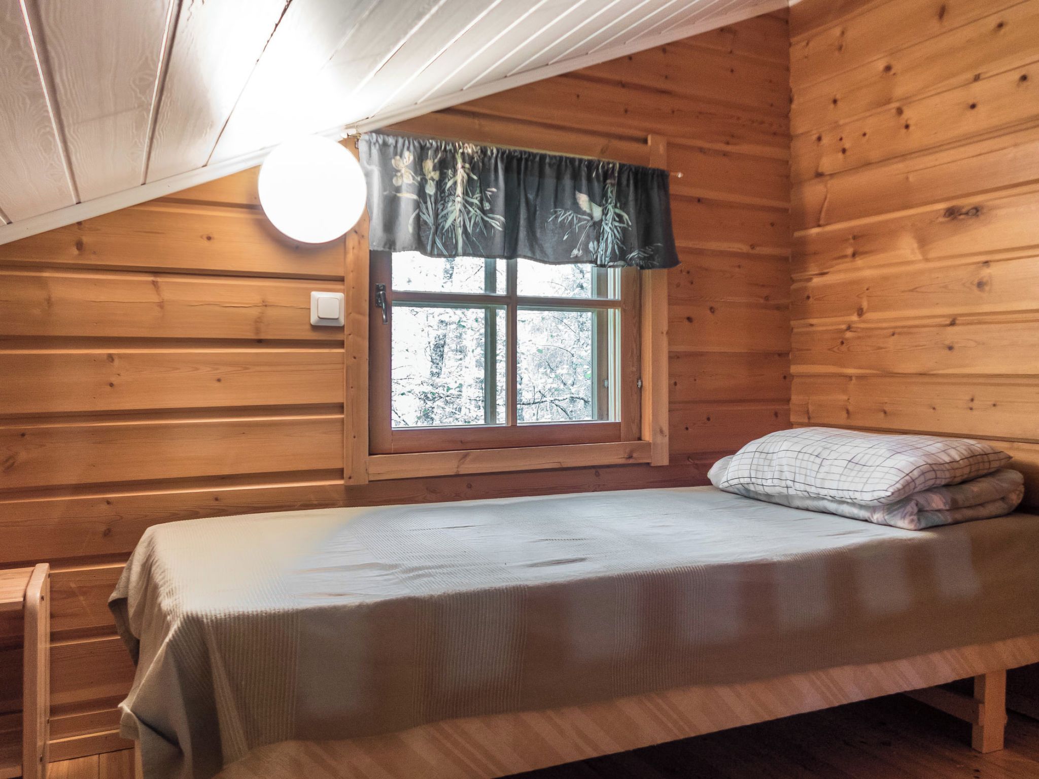 Una cama individual en una cabaña con ventana, paredes de madera y una lámpara.