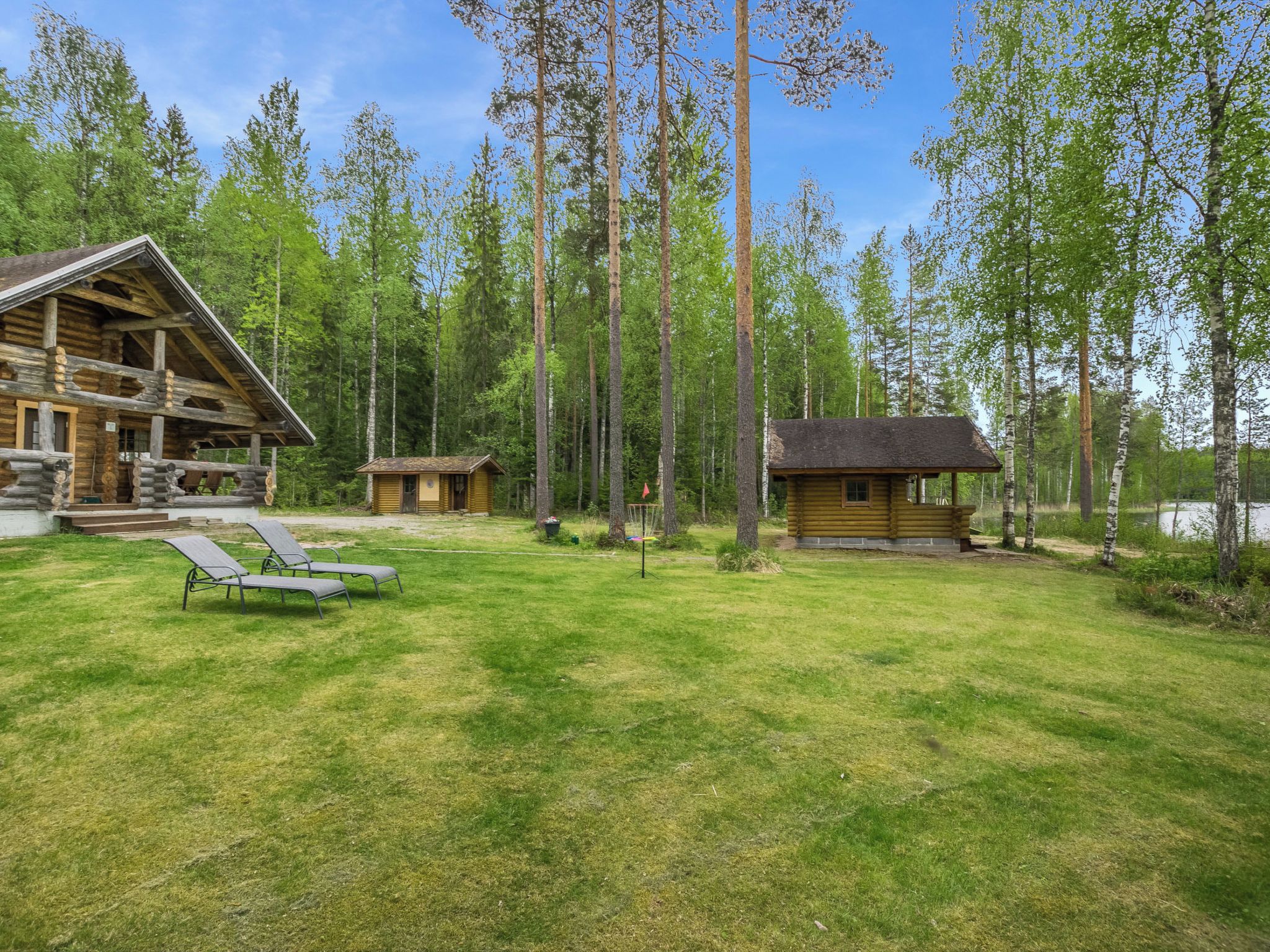 A log cabin with a porch and a small deck, surrounded by a grassy yard and tall trees.