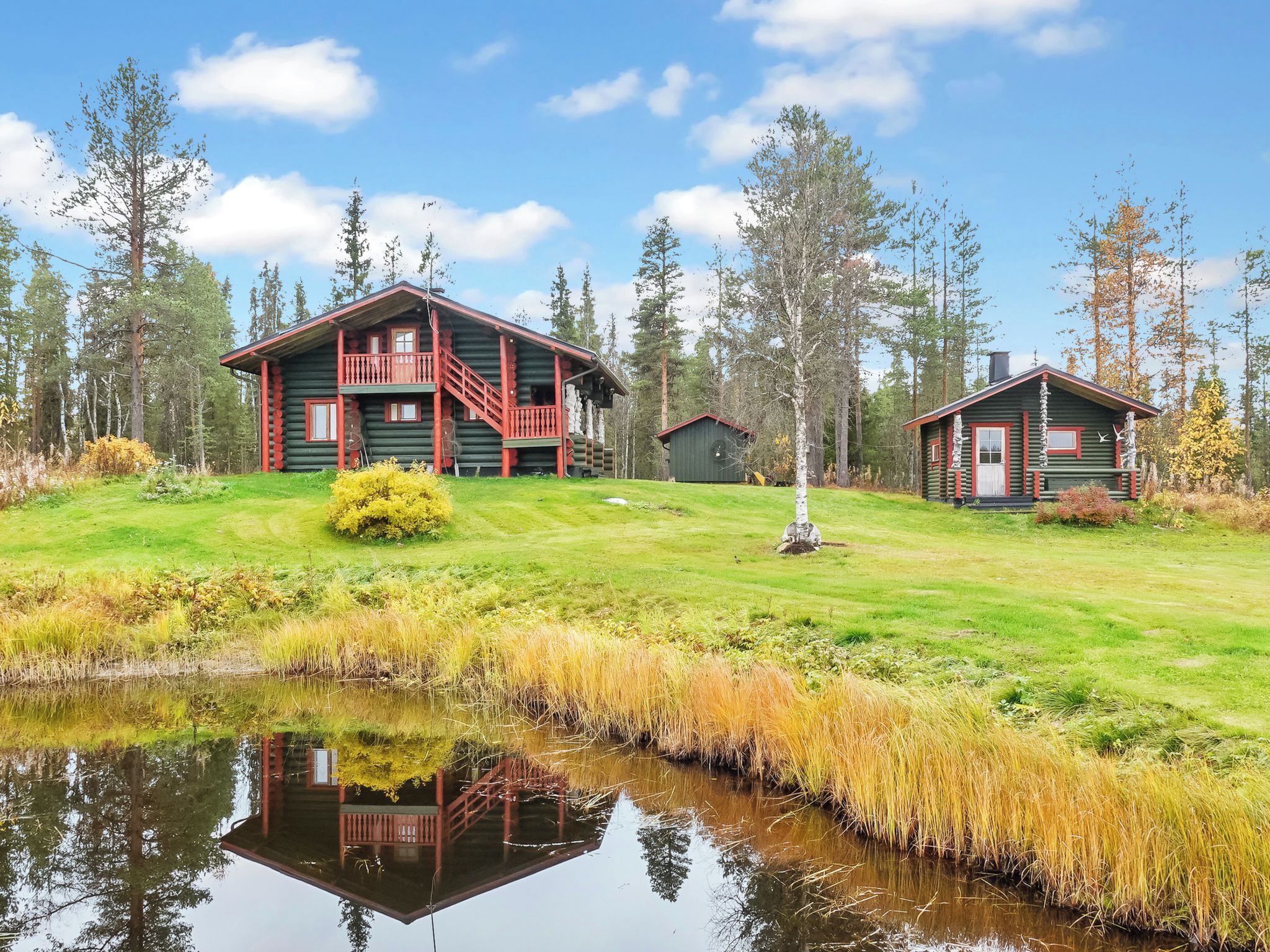 A log cabin with a balcony, a smaller cabin, and a pond in a forested area.
