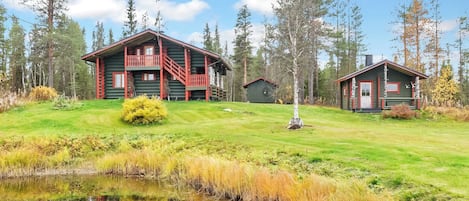 A log cabin with a balcony, a smaller cabin, and a pond in a forested area.