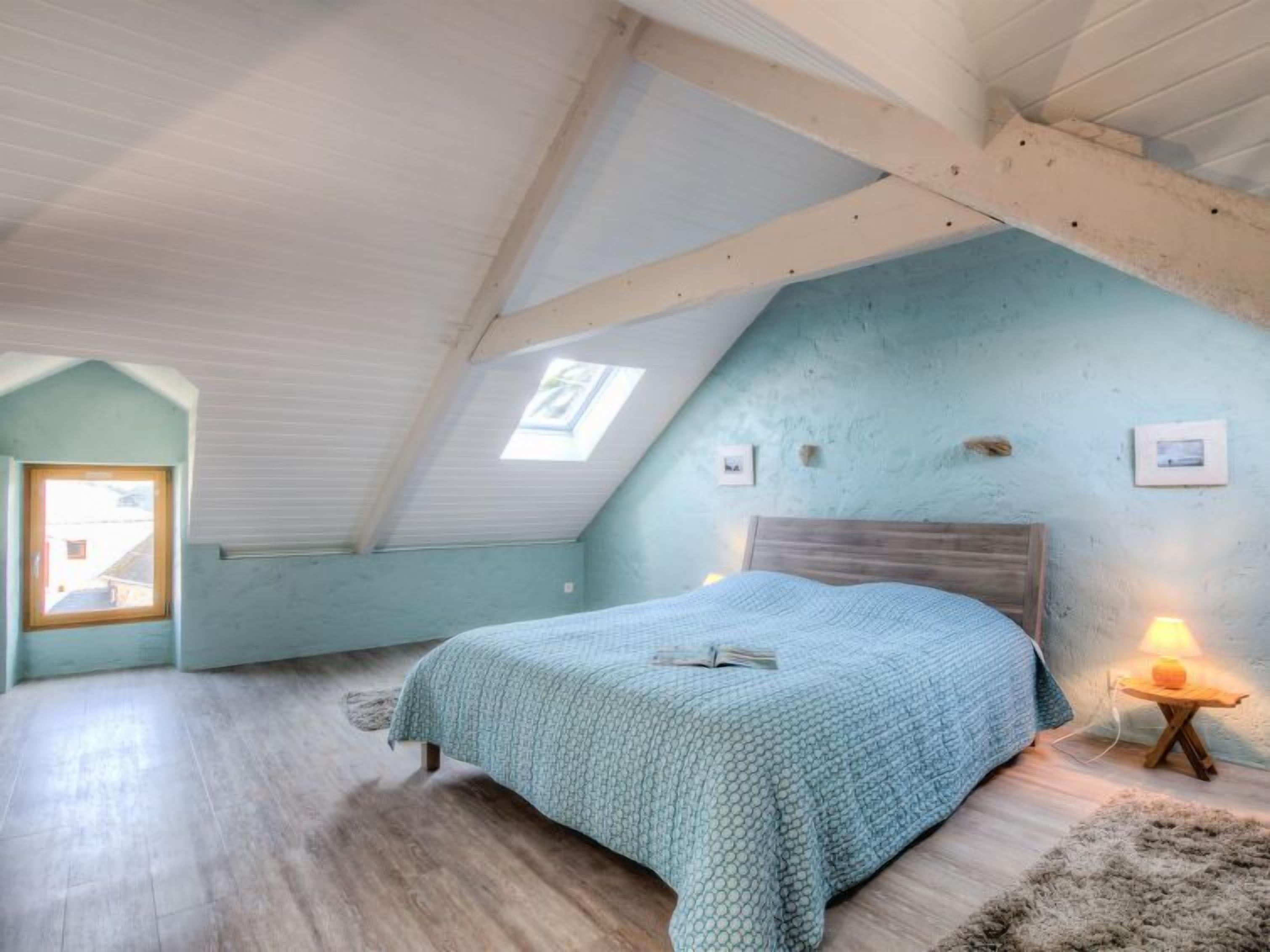 A bedroom with a sloped ceiling, a bed with a blue patterned bedspread, a small bedside table with a lamp, and a rustic wooden headboard.