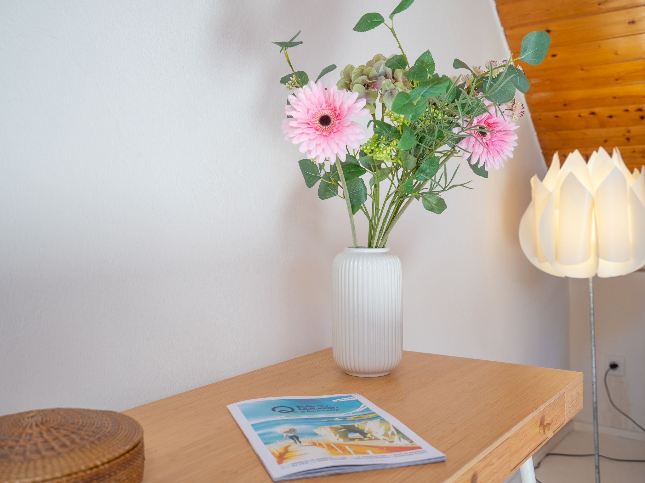 Eine weiße Vase mit rosa Blumen auf einem Holztisch, eine Zeitschrift und eine weiße Lampe.