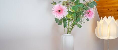 Eine weiße Vase mit rosa Blumen auf einem Holztisch, eine Zeitschrift und eine weiße Lampe.