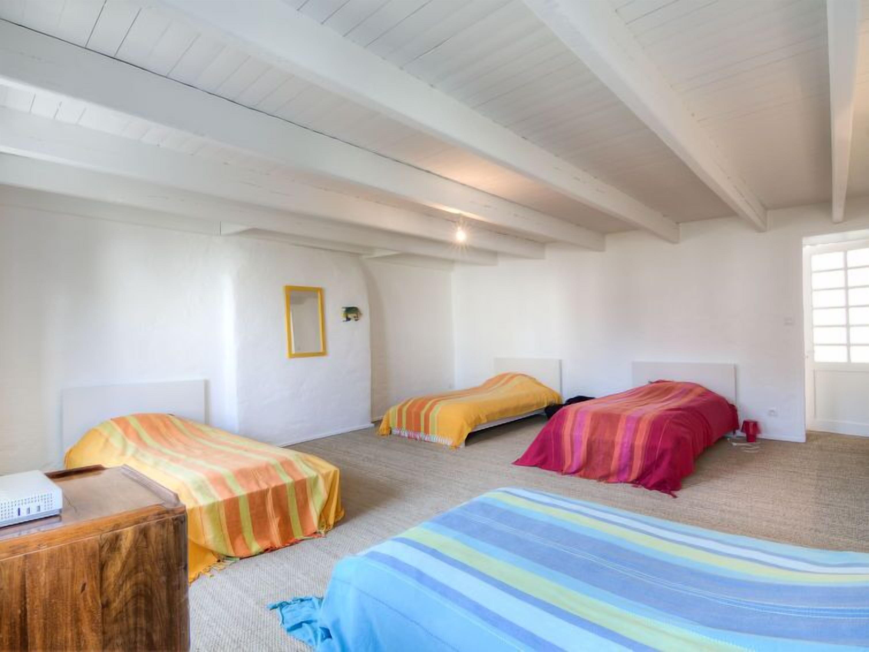 Een kamer met drie bedden, elk met een ander kleurend beddegoed. Er staat een houten nachtkastje met een apparaat erop, en een raam aan de rechterkant.