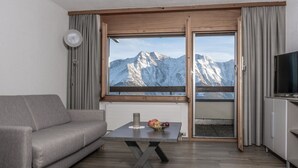 A modern living room with a sofa, a table with fruit, a TV, and a view of snow-capped mountains through large windows. - Zurschmitten 12 1. UG Süd by Interhome (Riederalp)