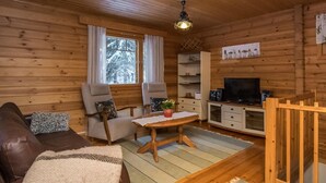 Ein gemütliches Blockhaus-Interieur mit Holzboden, einer Couch, zwei Sesseln, einem kleinen Tisch, einem Fernseher und einem Regal mit Dekorationsobjekten.