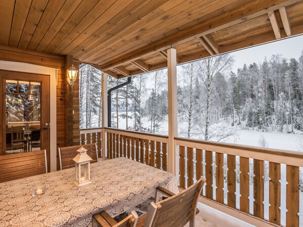 Una terraza de madera con una mesa, sillas, un farol y vistas a un bosque nevado.