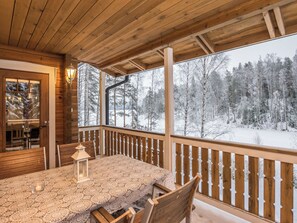 Een houten terras met een tafel, stoelen, een lantaarn en uitzicht op een besneeuwd bos.