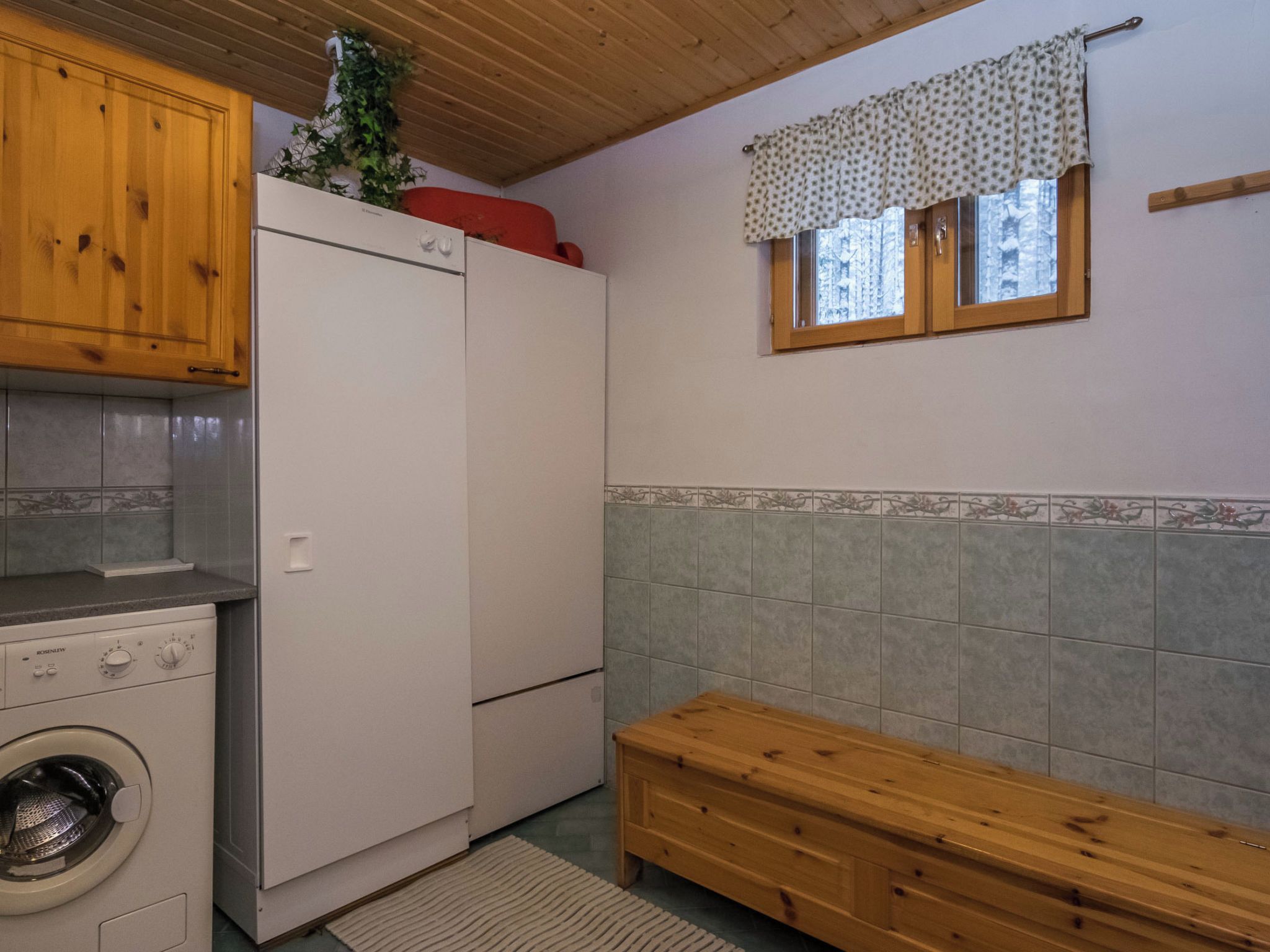 Une cuisine compacte équipée d’un lave-linge, d’un réfrigérateur, de meubles de rangement en bois et d’une fenêtre agrémentée d’une lambrequin à motifs.