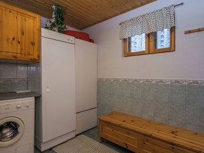Une cuisine compacte équipée d’un lave-linge, d’un réfrigérateur, de meubles de rangement en bois et d’une fenêtre agrémentée d’une lambrequin à motifs.
