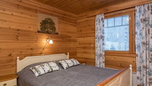 Ein Schlafzimmer einer Holzhütte mit einem Bett, einem Nachttisch, einer Wandlampe und einem Fenster mit floralen Vorhängen.