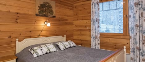 A wooden cabin bedroom with a bed, nightstands, a wall-mounted lamp, and a window with curtains.