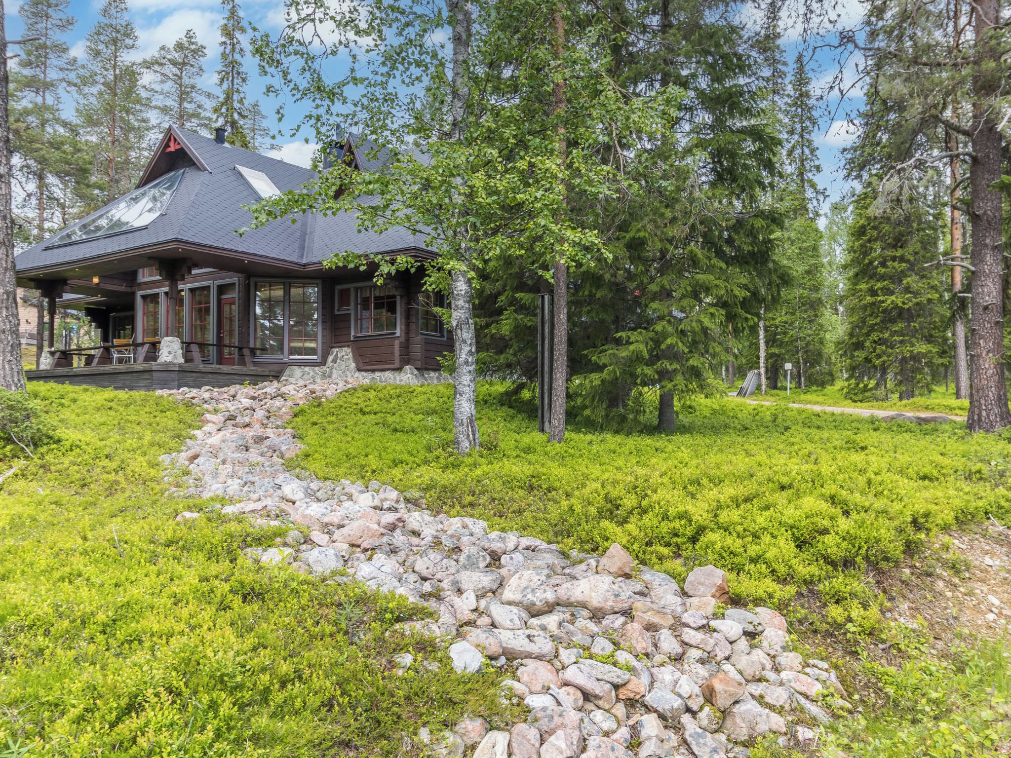Mökki, jonka edustalle johtaa kivipolku. Mökkiä ympäröi vehreä kasvillisuus ja korkeat puut.