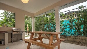 Ein überdachter Patio-Bereich mit einem Holztisch, einem Grill und einem Fliesenboden.