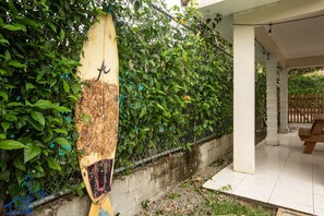 Relax on the covered patio, surrounded by lush greenery and a surfboard leaning against the fence.