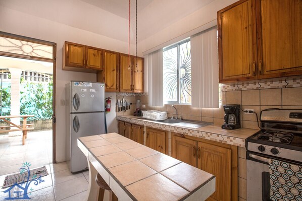 Kitchen features a stainless steel refrigerator, gas stove, sink, and coffee maker, with a window and wooden bench nearby.
