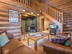 Interior de una cabaña de troncos con chimenea, escalera de madera, sofás de cuero y televisor.