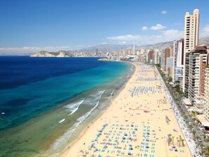 A coastal cityscape with a wide sandy beach lined with tall buildings, a clear blue sea, and a mountainous backdrop.