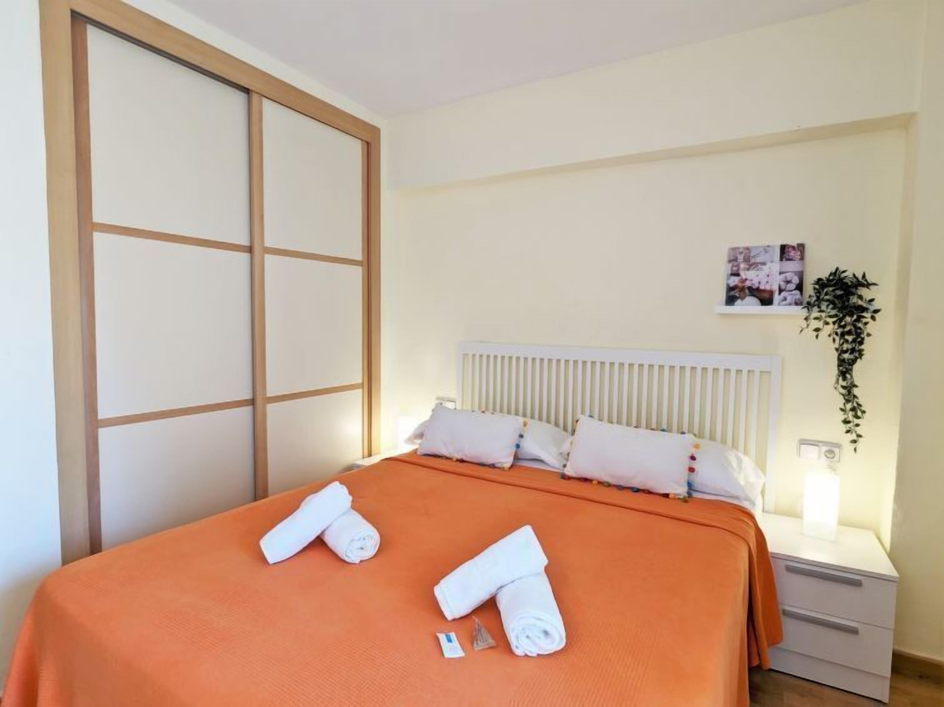 Une chambre d’hôtel avec un lit, des oreillers blancs, une couvre-lit orange, une table de chevet blanche avec une lampe et une plante fixée au mur.