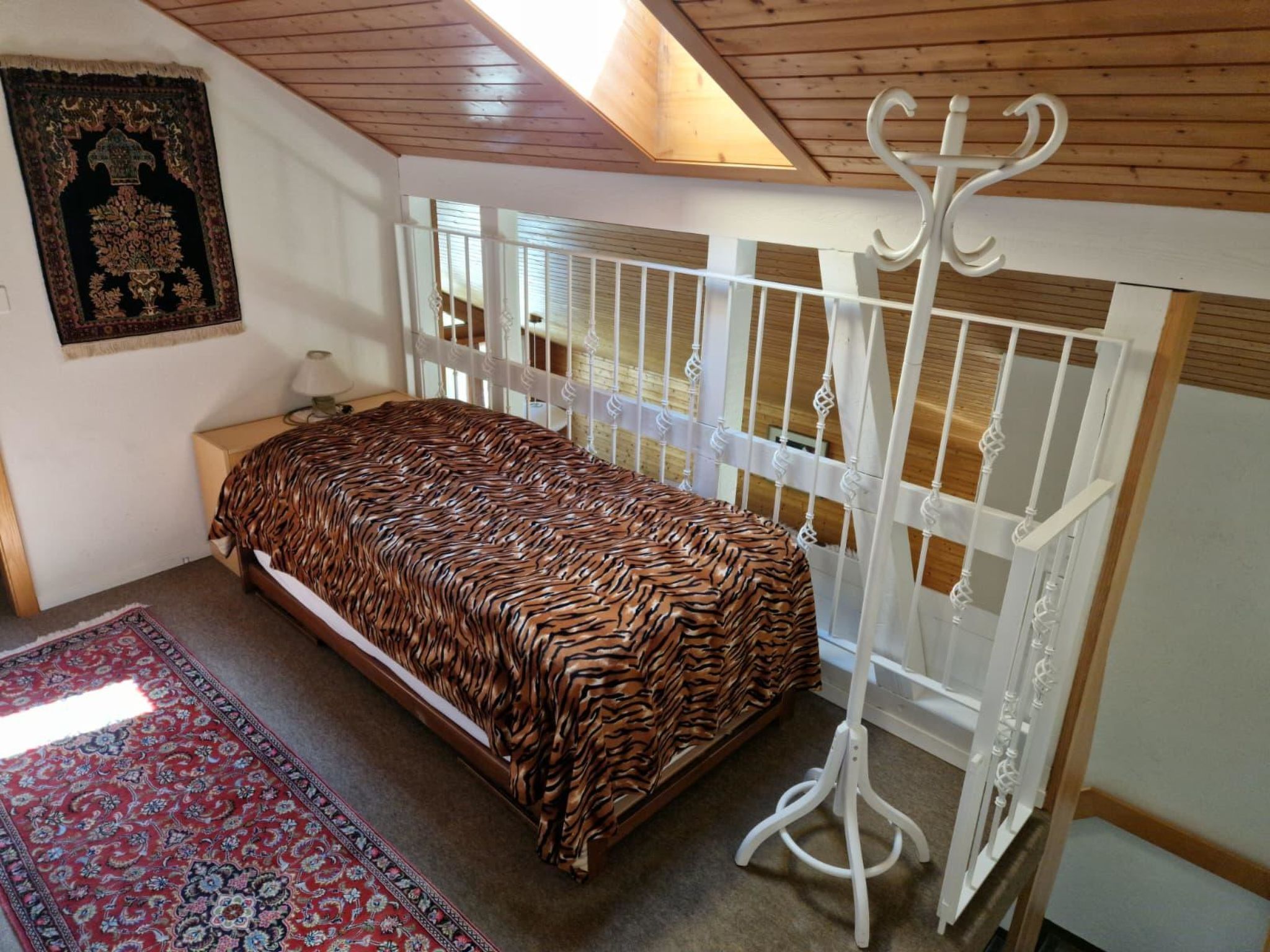 Une chambre à coucher avec un lit, un cadre de lit en métal blanc, un tapis rouge à motifs et un plafond en bois.