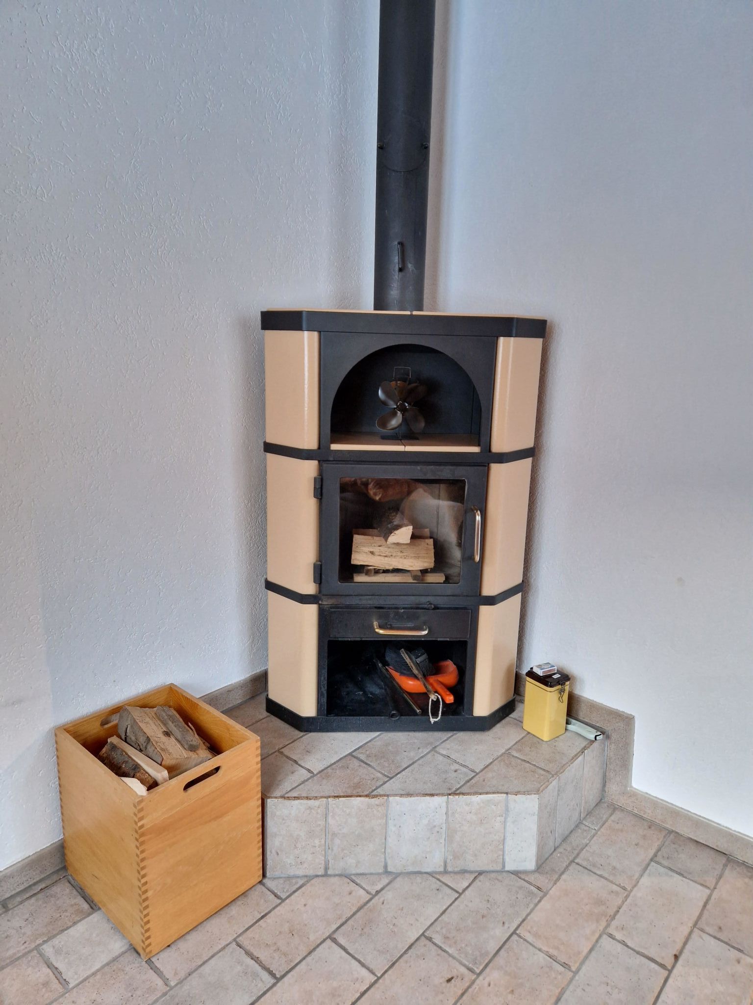 Una chimenea en esquina con base de piedra, una caja de madera con leña y un recipiente amarillo.