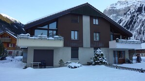 Una casa de dos pisos con un balcón, un garaje y una montaña nevada al fondo.