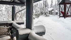 A wooden cabin with a log fireplace, a hanging lantern, and a snowy landscape with trees and a cabin in the background.