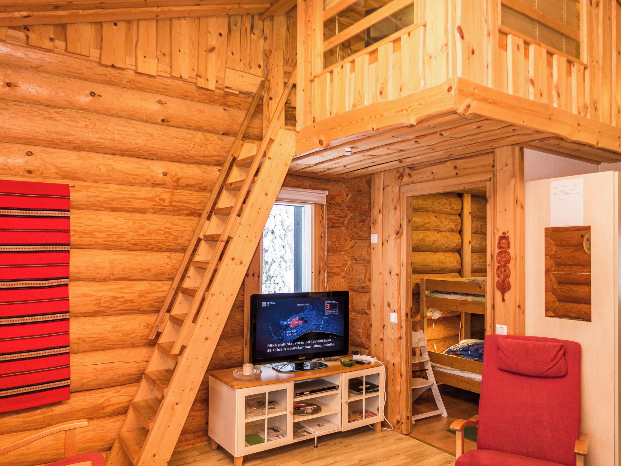 Een houten blokhut van binnen met een televisie, een rode stoel en een houten ladder.