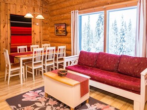 A cozy cabin interior with a wooden wall, a dining table with chairs, a sofa, and a large window with a snowy view.