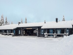A log cabin with a snow-covered roof, a sign reading 'RUKAN LEPLINNA', and a wooden fence in front.