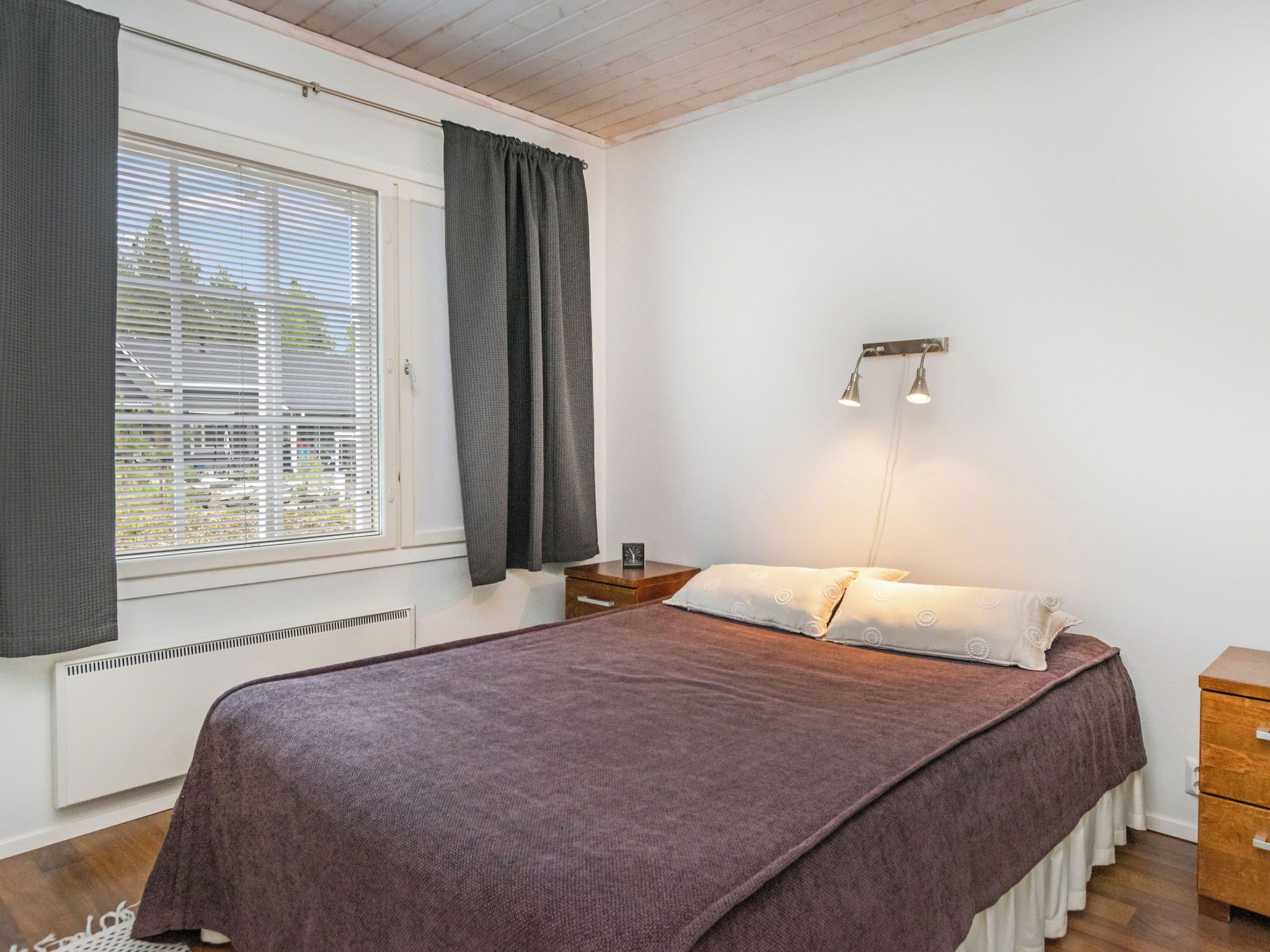 A bedroom with a bed, a nightstand, a wall-mounted light fixture, and a window with blinds.
