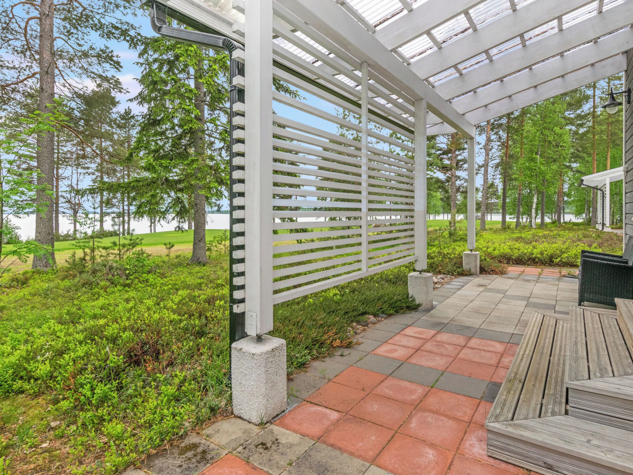 Katettu ulkotila, jossa on pergola, kivetty kävelyreitti ja näkymä järvelle.