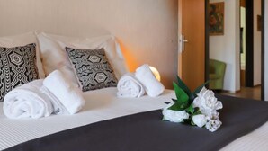Una cama bien hecha con toallas blancas enrolladas, almohadas estampadas y un ramo de flores blancas sobre un edredón oscuro.