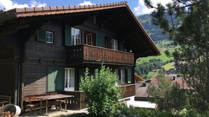 A two-story wooden house with a balcony, a wooden table and bench outside, and a view of a mountainous landscape.