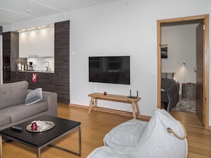 Uma sala de estar moderna com sofá, mesa de centro e TV. Há um banco de madeira e uma pequena mesa com um vaso. Uma cozinha é visível ao fundo.